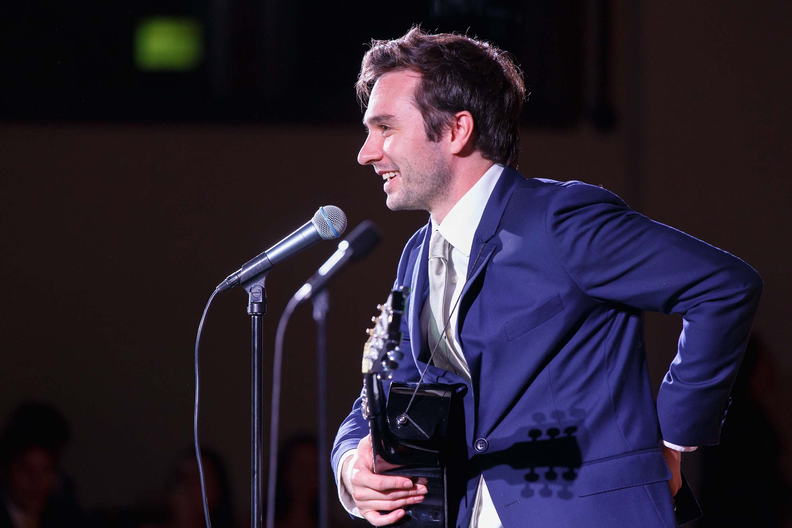 A student wearing a blue suit smiling at a microphone, holding a guitar.
