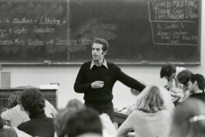 A professor in a black sweater stands in front of a chalk board teaching students.