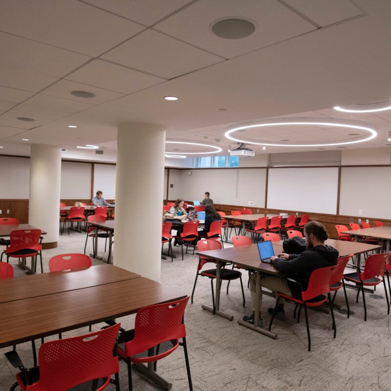 Students engaged in various activities sit in a large open space with several long tables with chairs and overhead lighting.