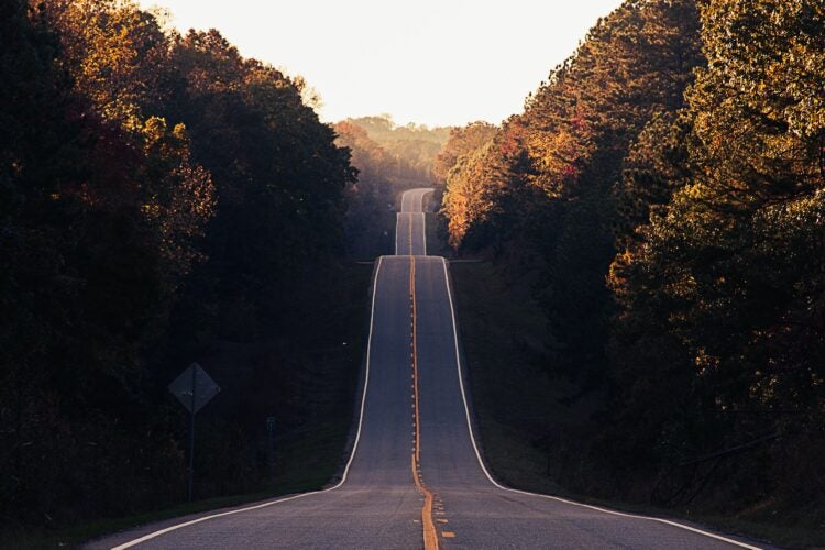 View of a tree-lined two lane highway with no cars on the road. The road had many dips up and down and curves off into the distance.