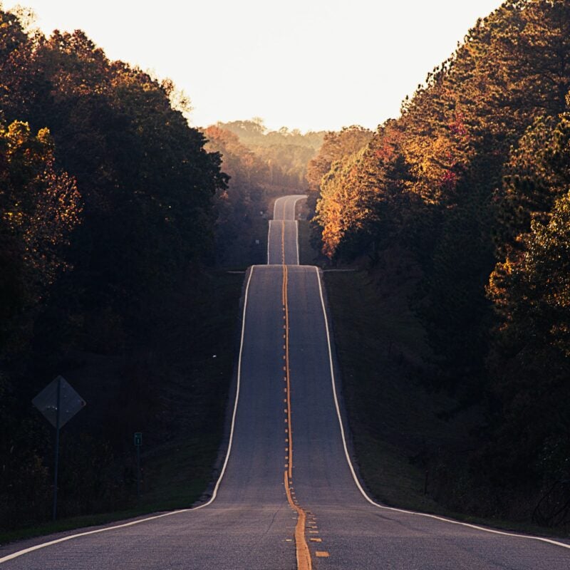 View of a tree-lined two lane highway with no cars on the road. The road had many dips up and down and curves off into the distance.