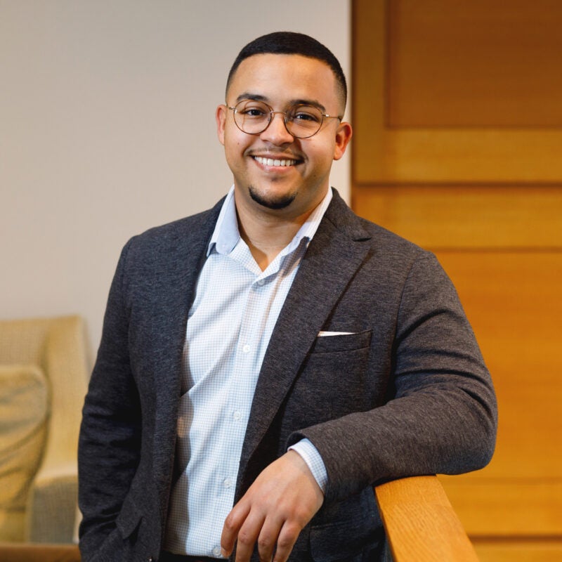 Portrait of Carlos Gonzalez Sierra wearing a grey blazer leaning against a railing.