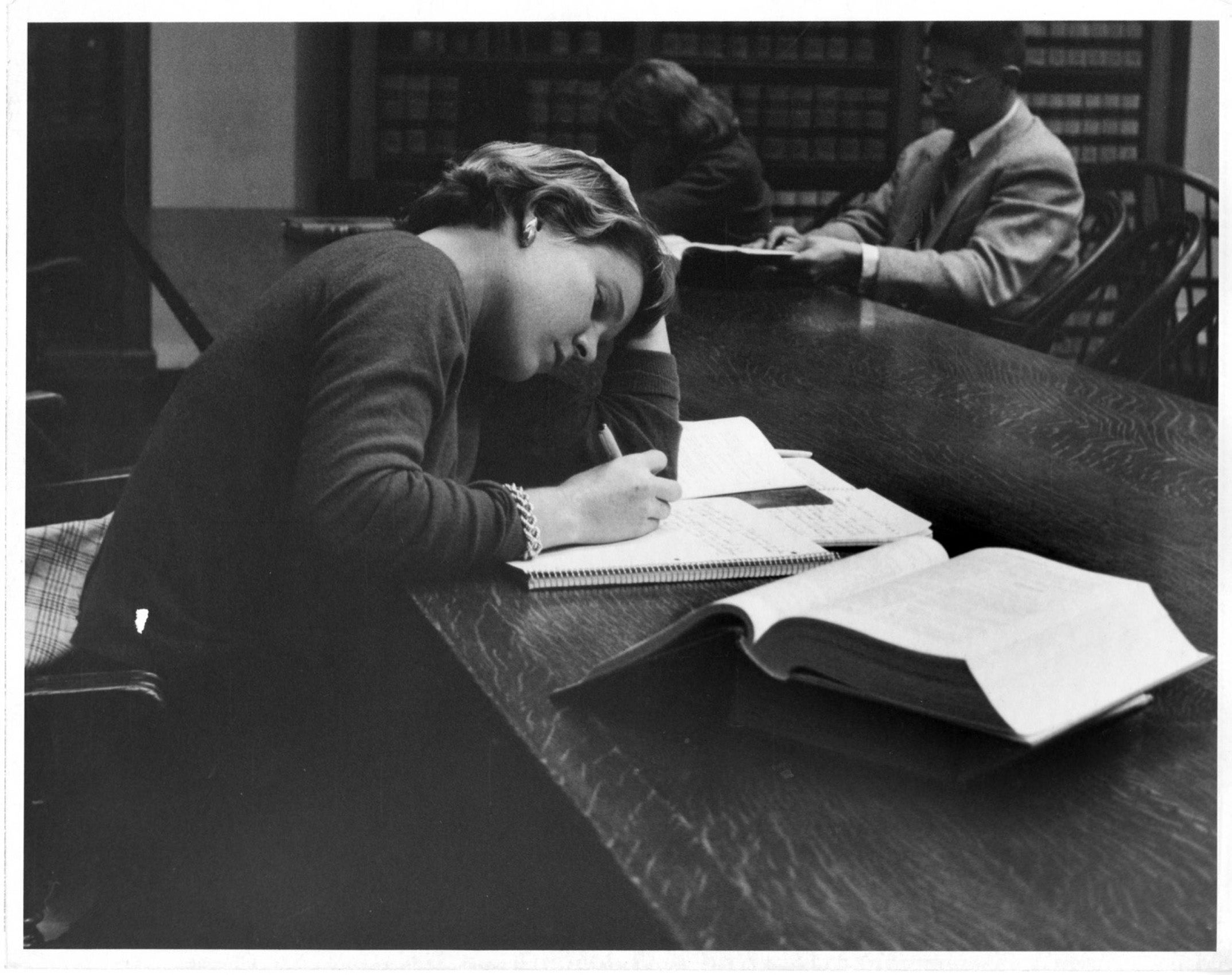 A black and white photo of a woman in the library leaning on her elbow as she studies.