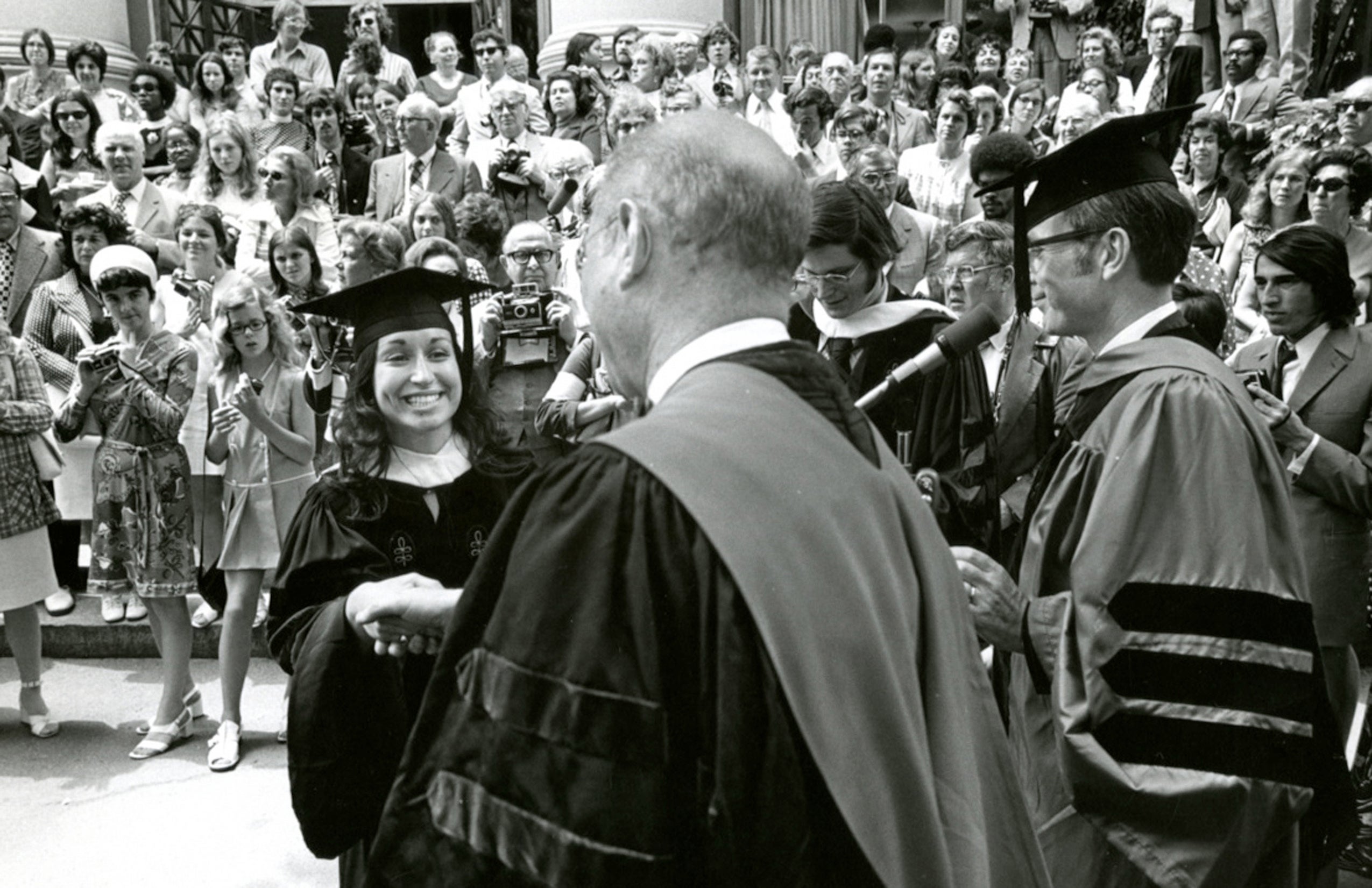 A woman wearing commencement regalia shakes the hand of the dean as a large crowd looks on