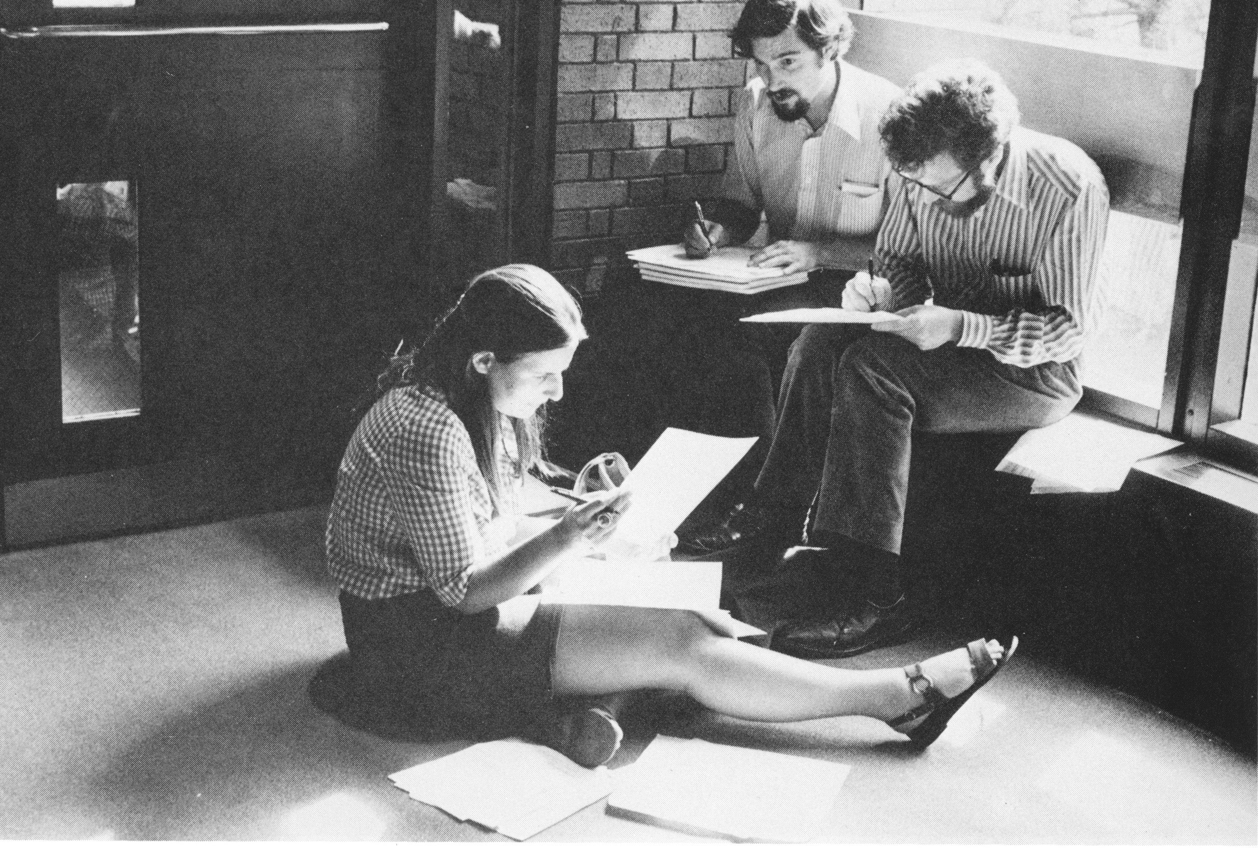 Two men sitting on and interior window ledge and one women sitting on the ground going over papers