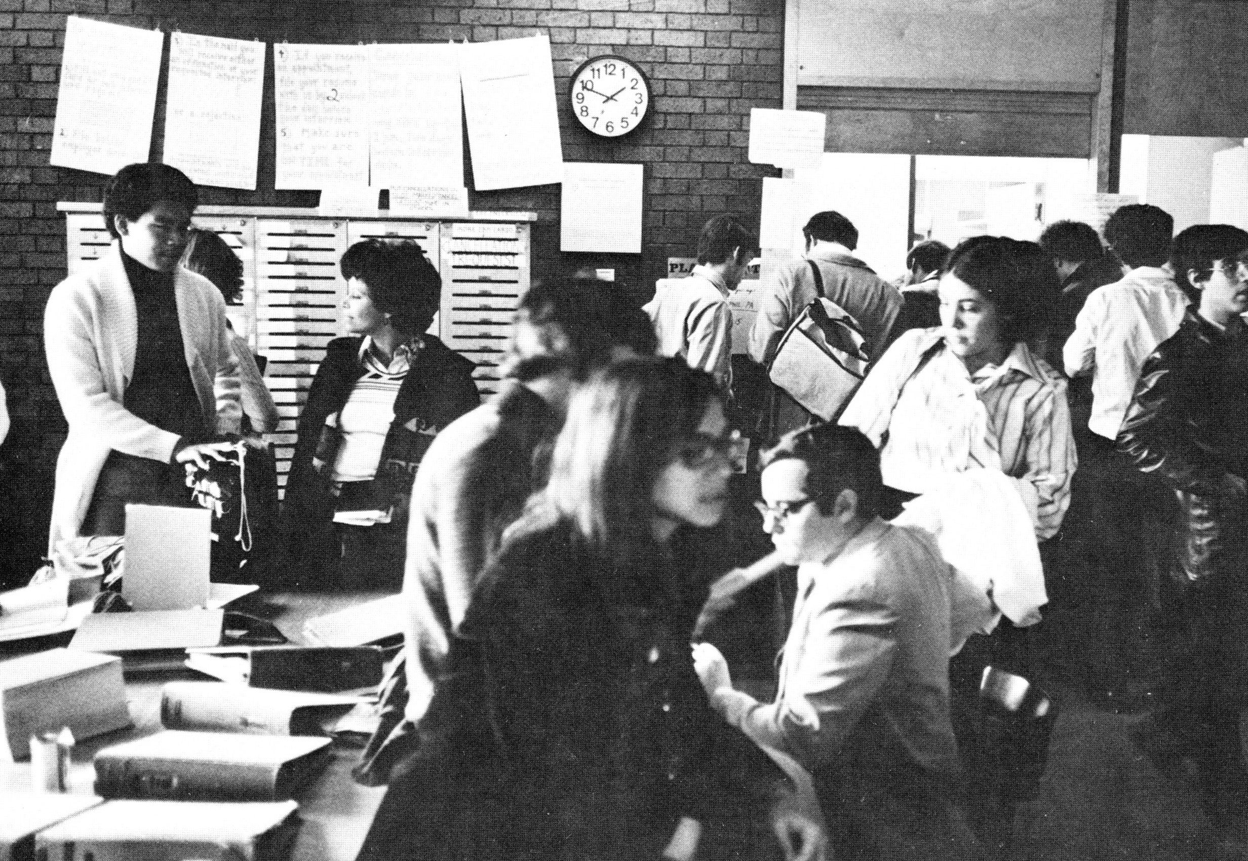 A crowd of men and women move around in a space with a table of notebooks