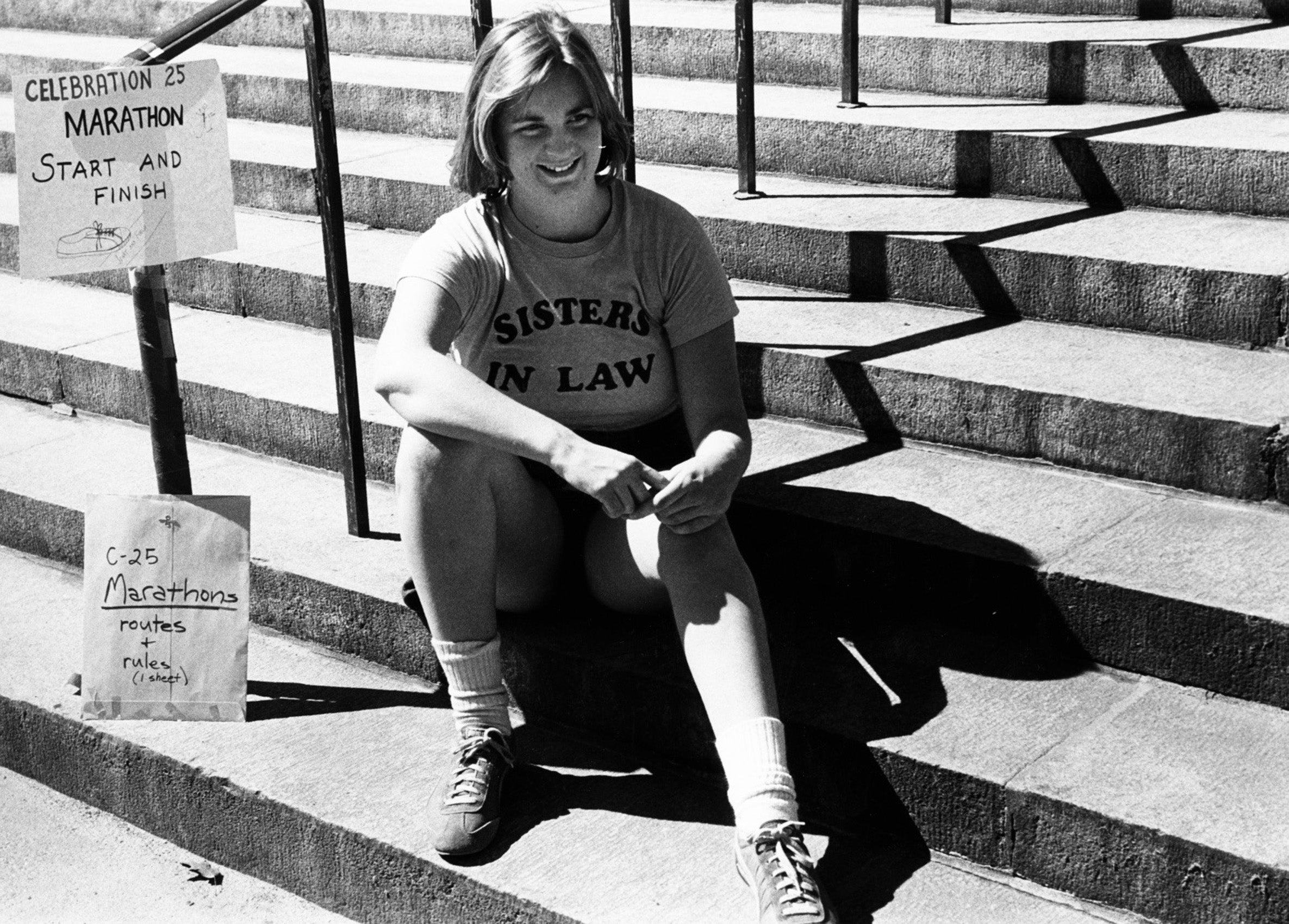 A woman sits on the stairs wearing the T-Shirt Sisters in Law with a sign Celebration 25 Marathon next to her on a railing