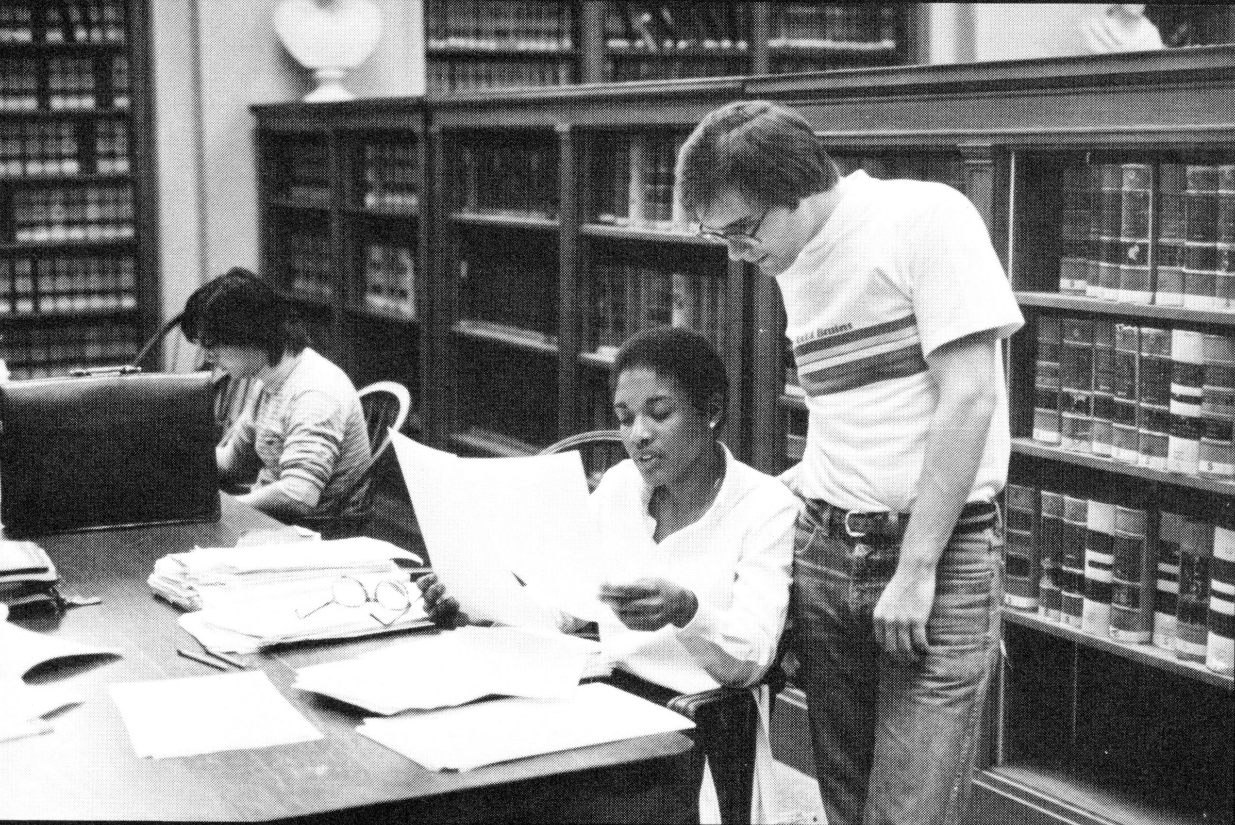 A women sitting at a table in the library shows a paper in her hands to a male students