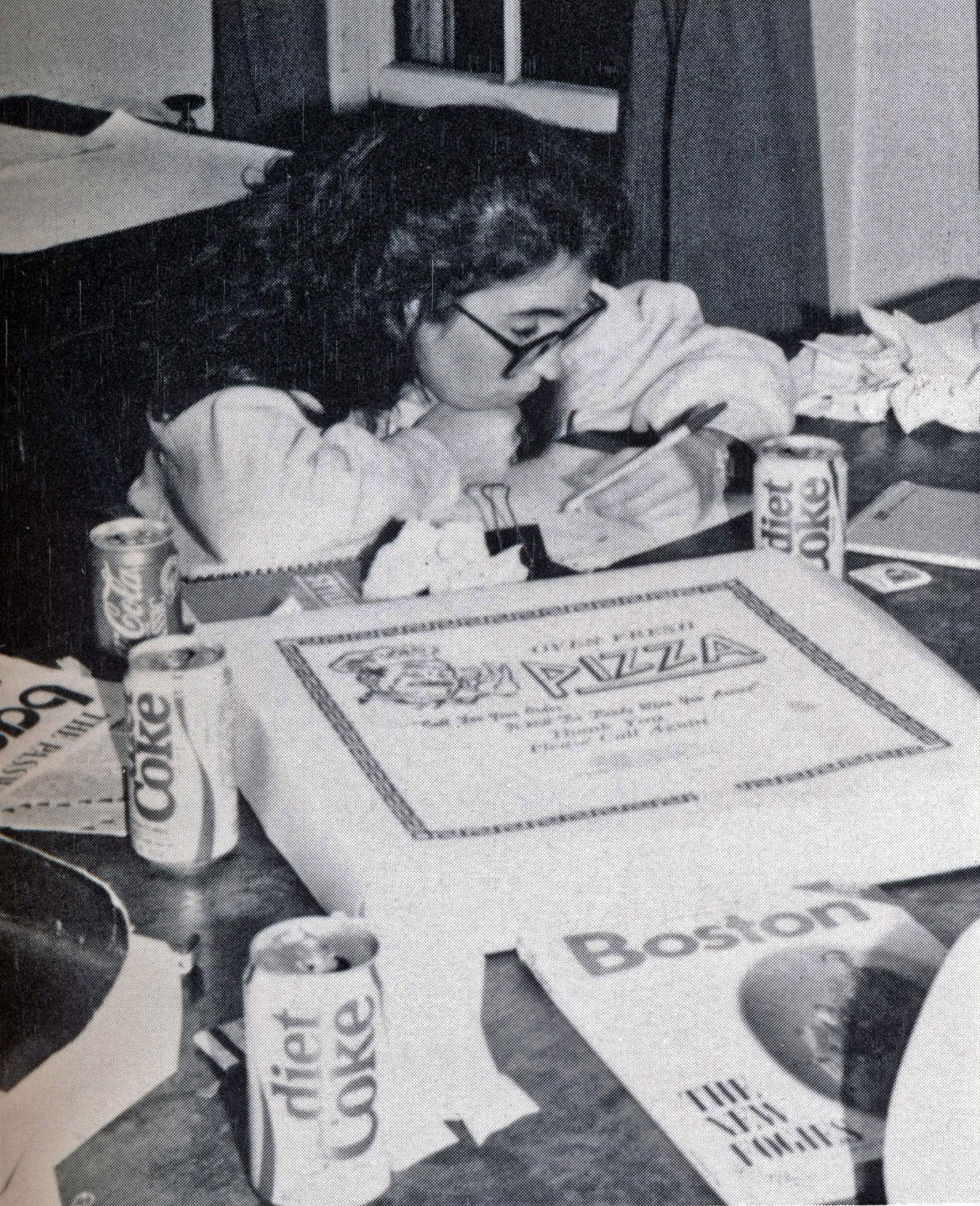 A woman working on a paper with a table with diet cokes and a pizza box