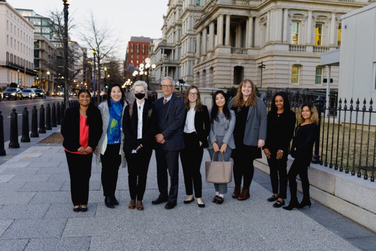 Nine individuals pose for a photo on the sidewalk of a street