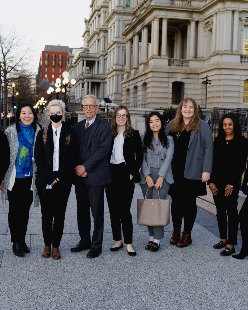 Nine individuals pose for a photo on the sidewalk of a street
