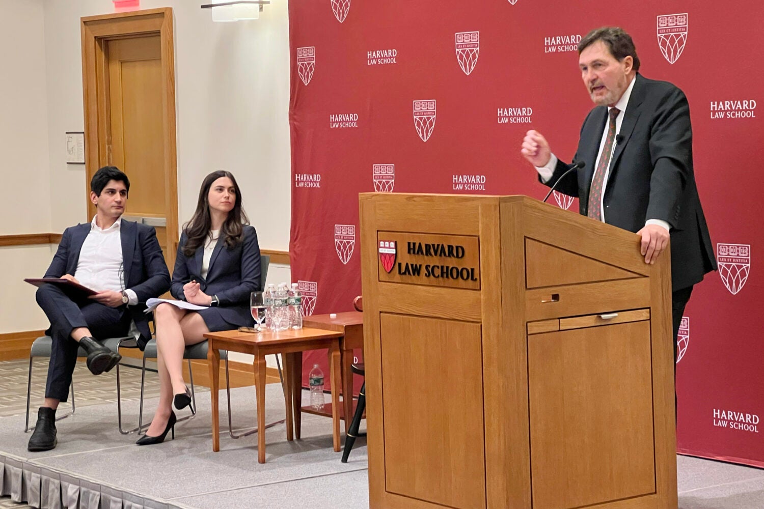 A man speaking at a podium with two people sitting to his right ins chairs