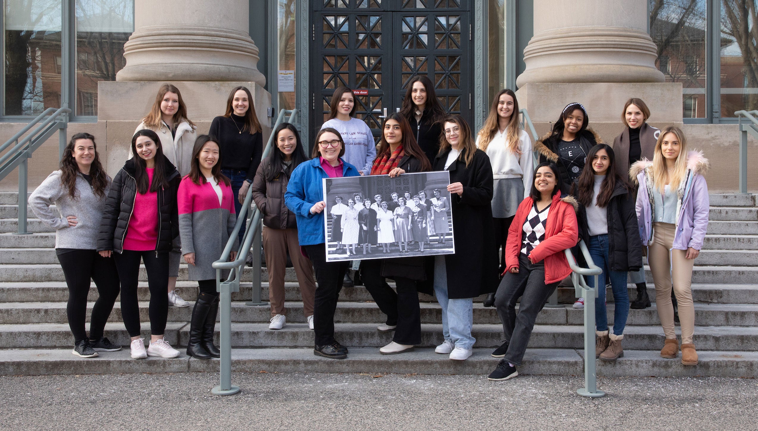 A group of 19 women pose on the steps of a building holding an archival photo of women posing on the same steps