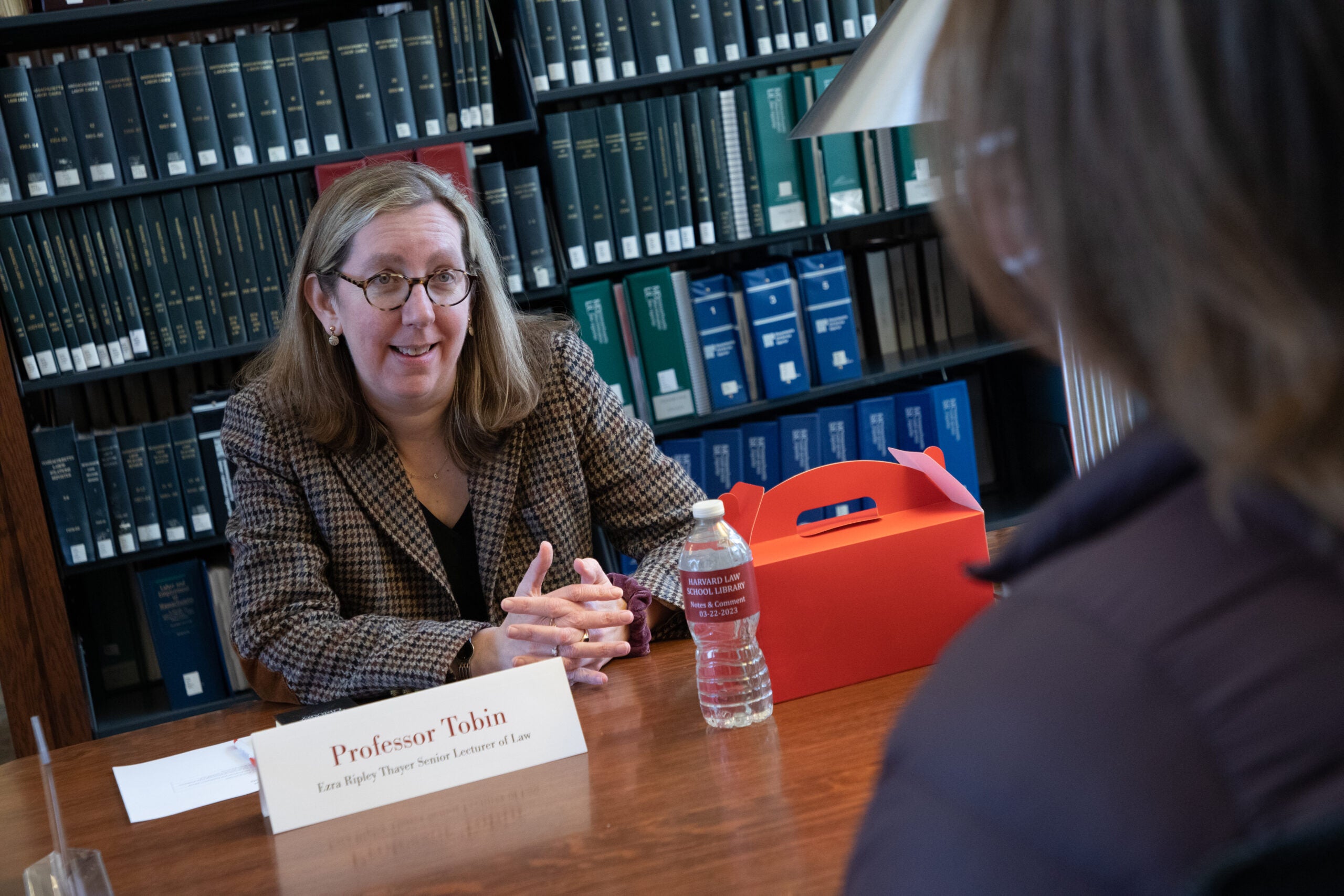 A woman sitting at a table in a library speaking with another woman