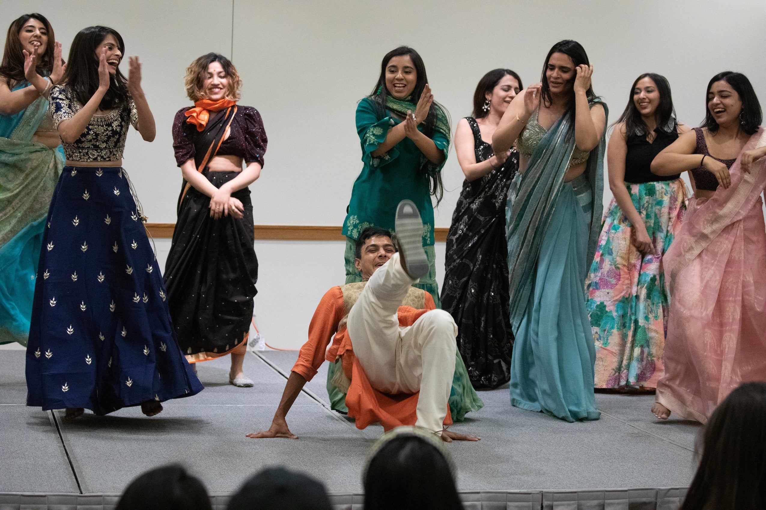 A man dances on the floor as a group of women dance behind him on a stage