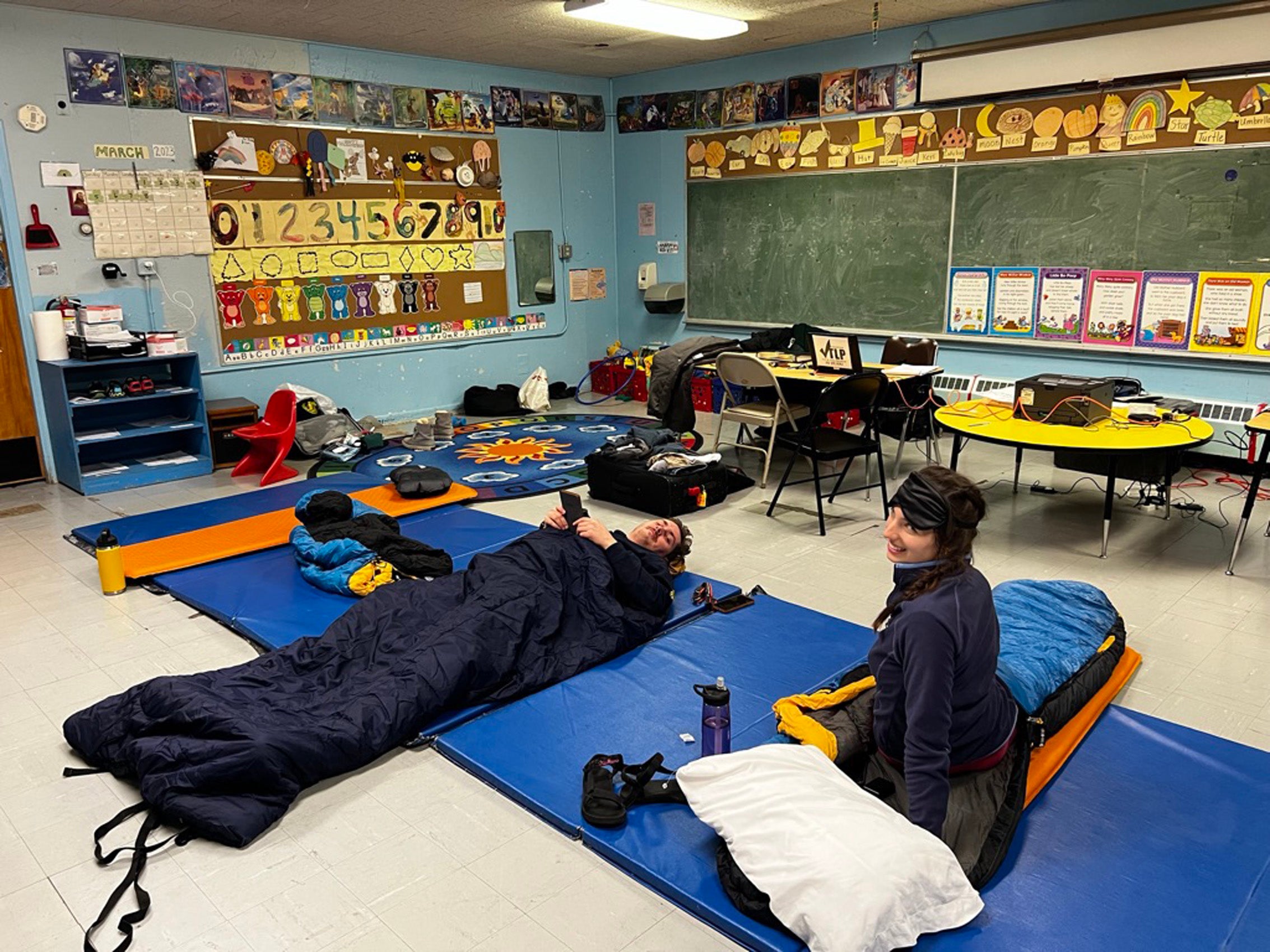 Two people sleeping in sleeping bags on the floor of a classroom