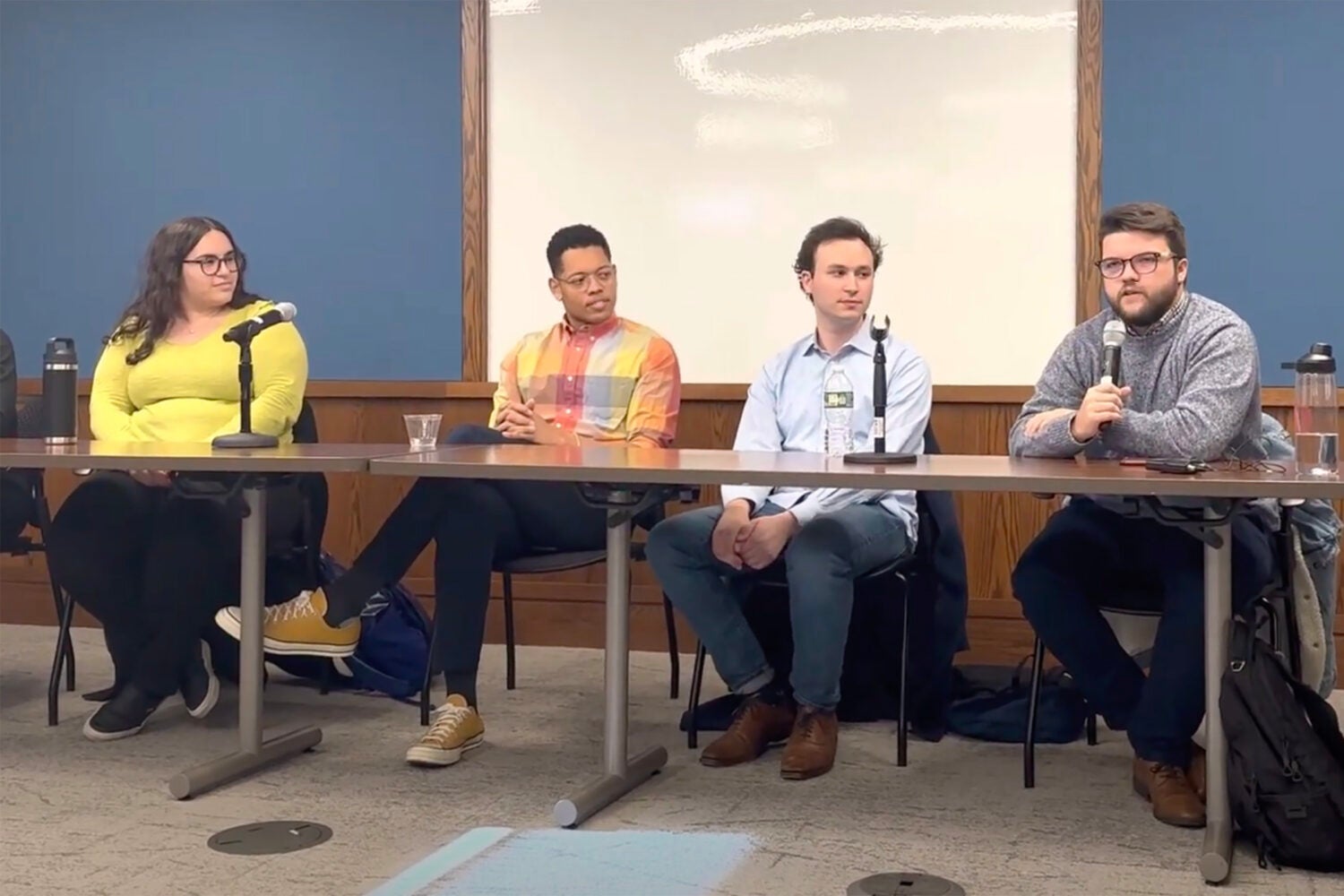 Four students seated at a table, one addressing an audience while holding a microphone.