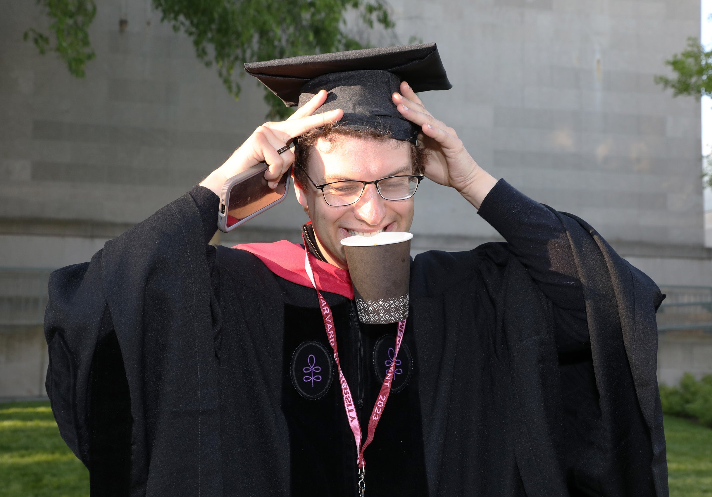 Graduate adjusts his cap while holding a paper cup in his teeth