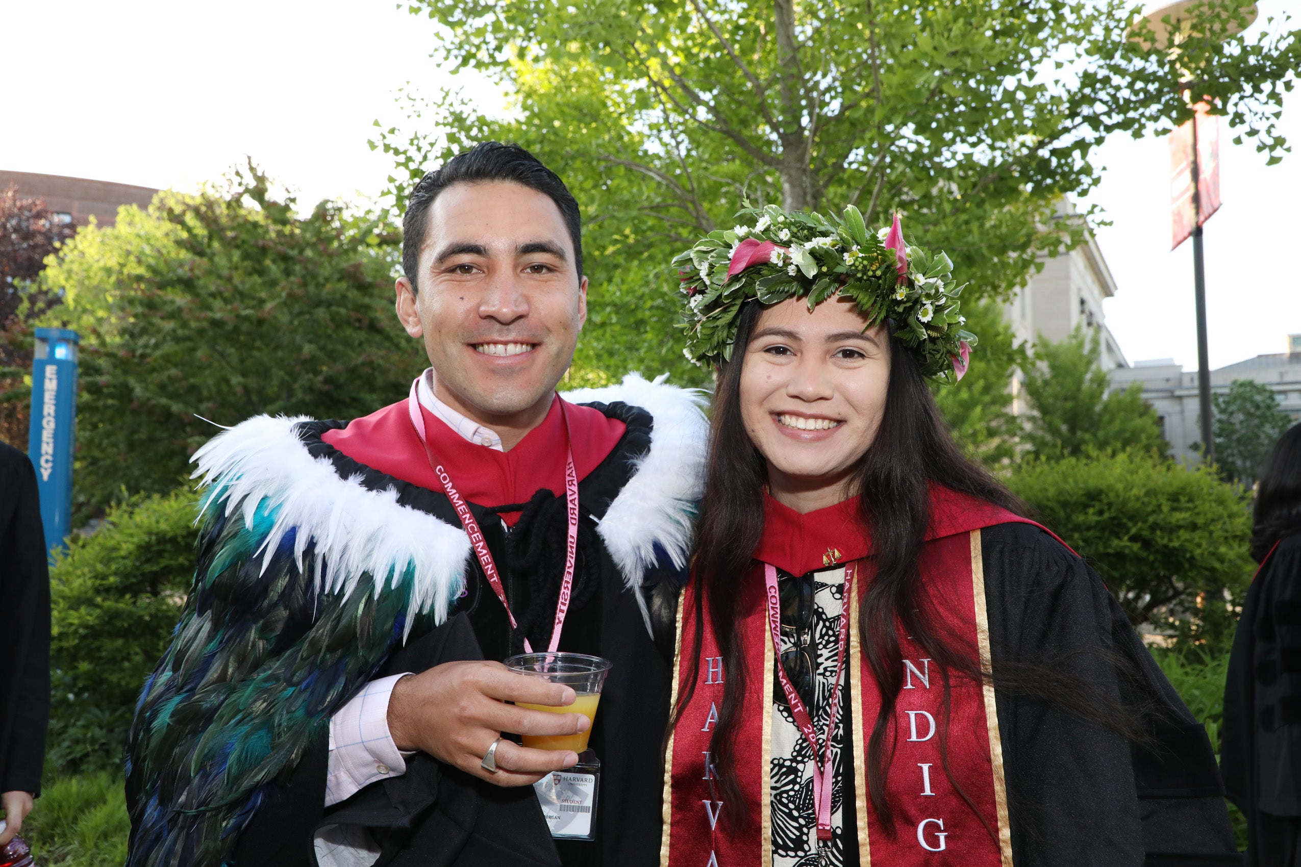 Male graduate wearing feather cape and female graduate wearing flower crown pose in their regalia