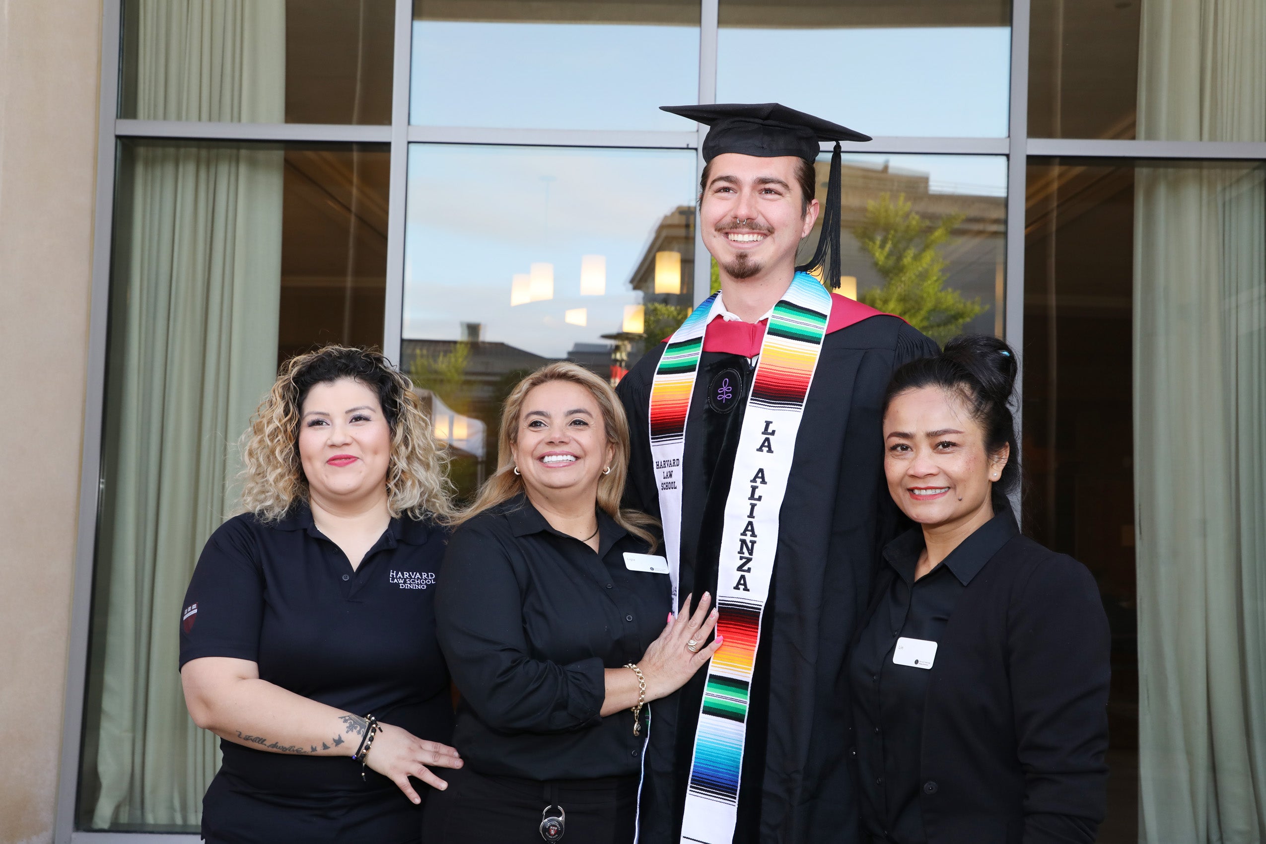 Tall male graduate wearing La Alianza sash poses with dining staff members