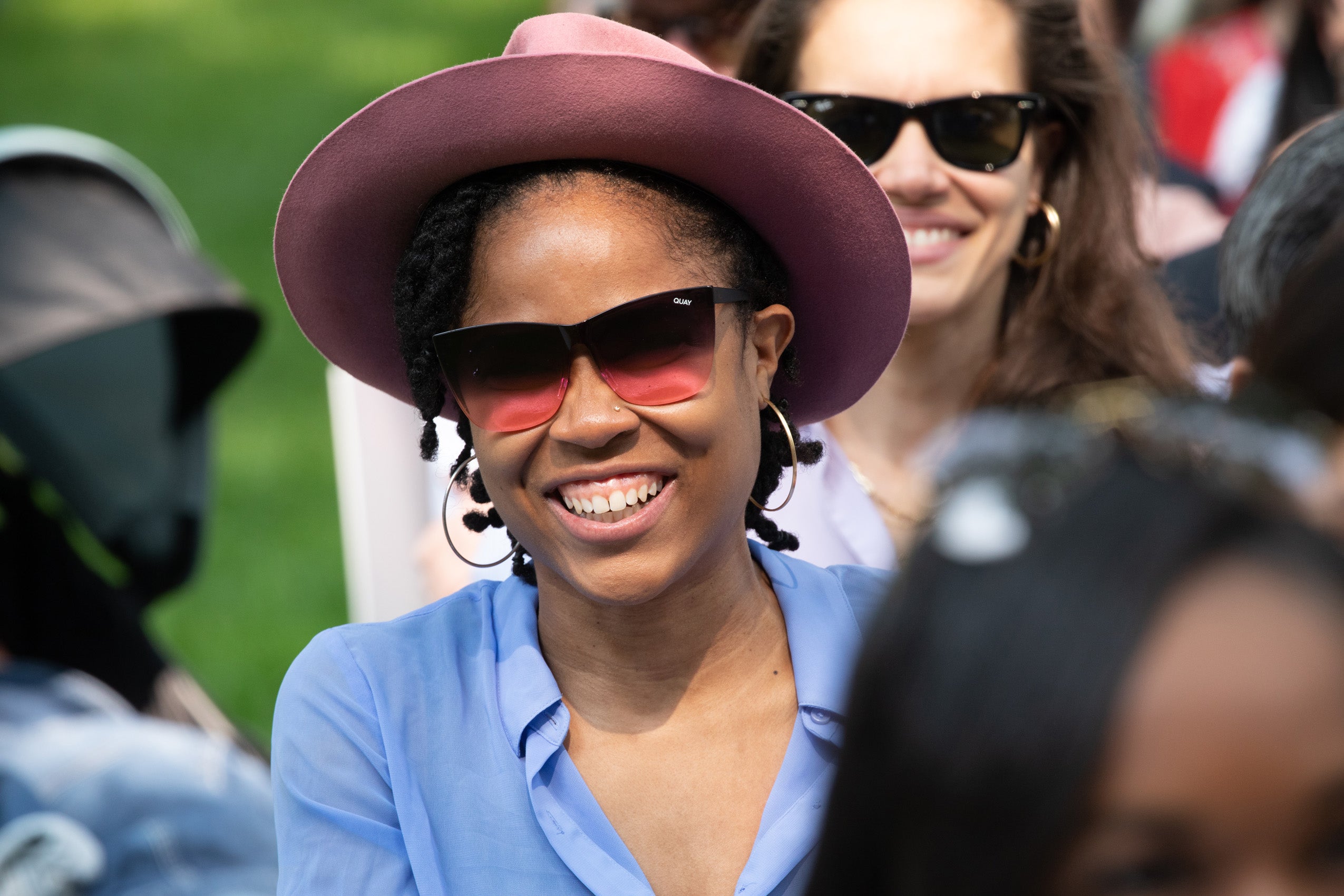 Woman in gradient sunglasses and mauve hat smiles in crowd