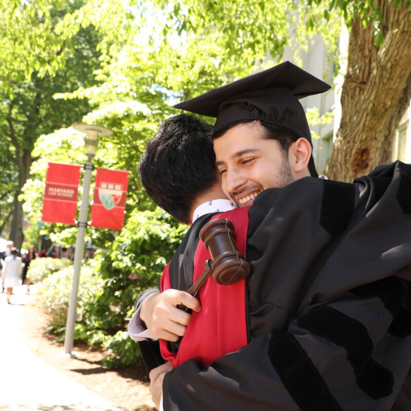 Two graduates wearing caps and gowns hug on a pathway on campus