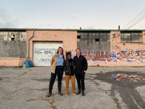 Three people stand in a dilapidated lot