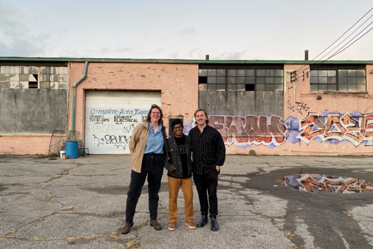 Three people stand in a dilapidated lot