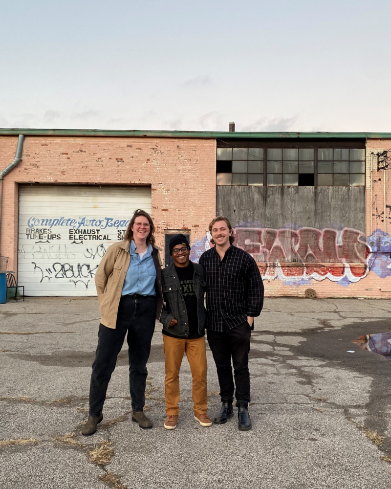 Three people stand in a dilapidated lot