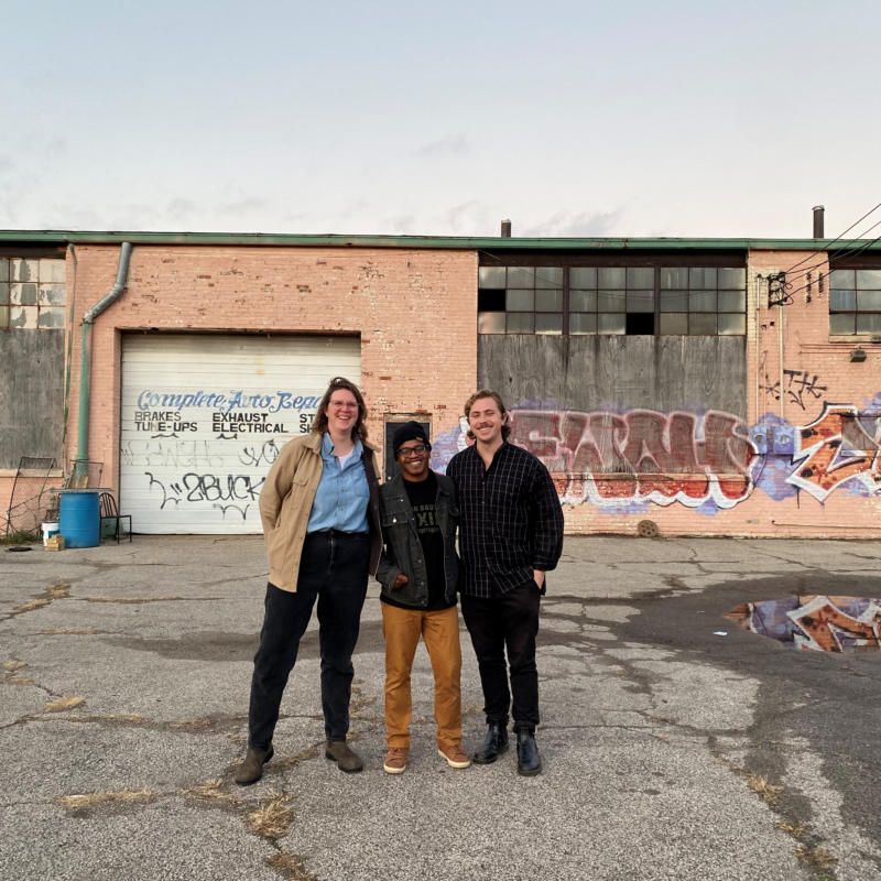 Three people stand in a dilapidated lot