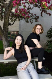 Two students sitting on a table wearing sitting under a pink flowering tree showing off their tattoos