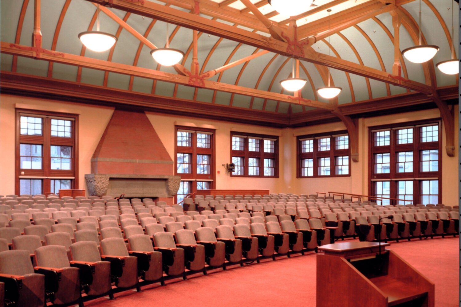 An overview shot of a large room with lots of large windows and rows of chairs. A brick fireplace with stone carvings sits in the background.