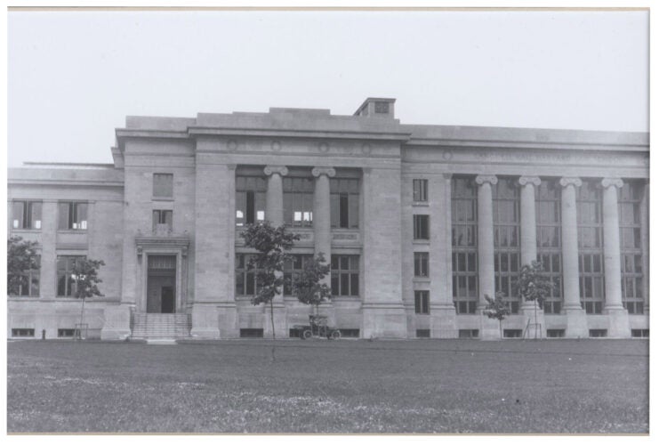 A large building on a grassy field with small trees and a single car