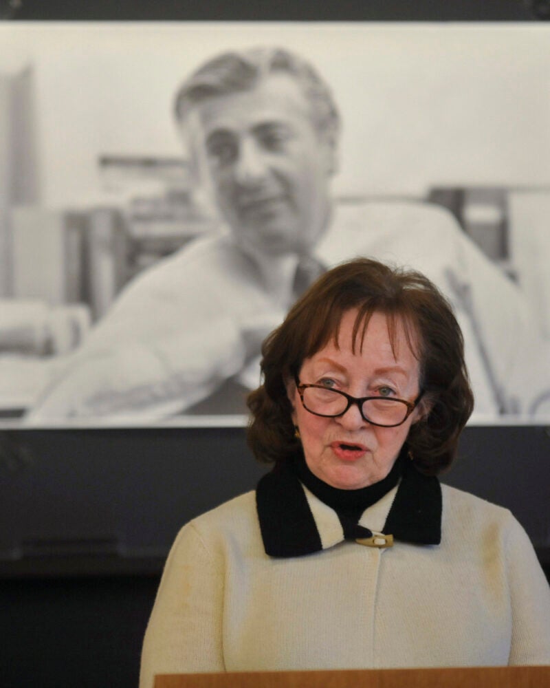 A woman speaking at a podium in front of a black and whit photograph of a man