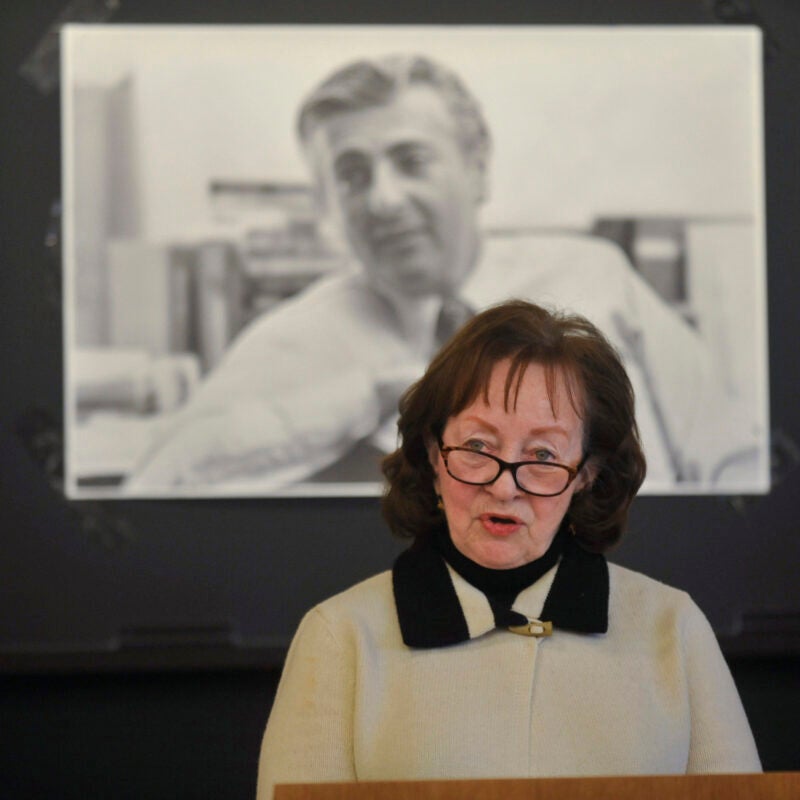A woman speaking at a podium in front of a black and whit photograph of a man