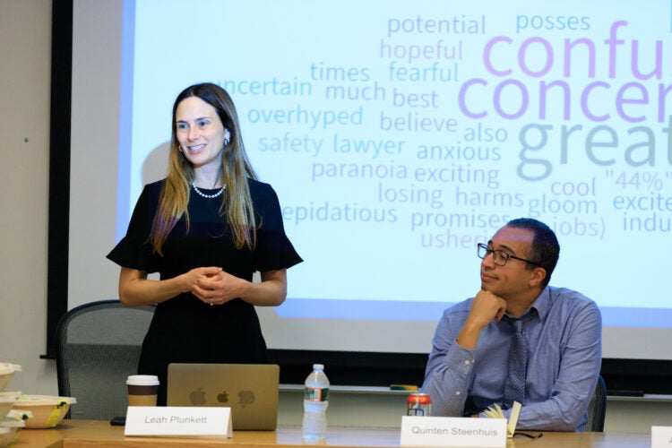 a woman stands in front of a projector screen, a man sits next to her