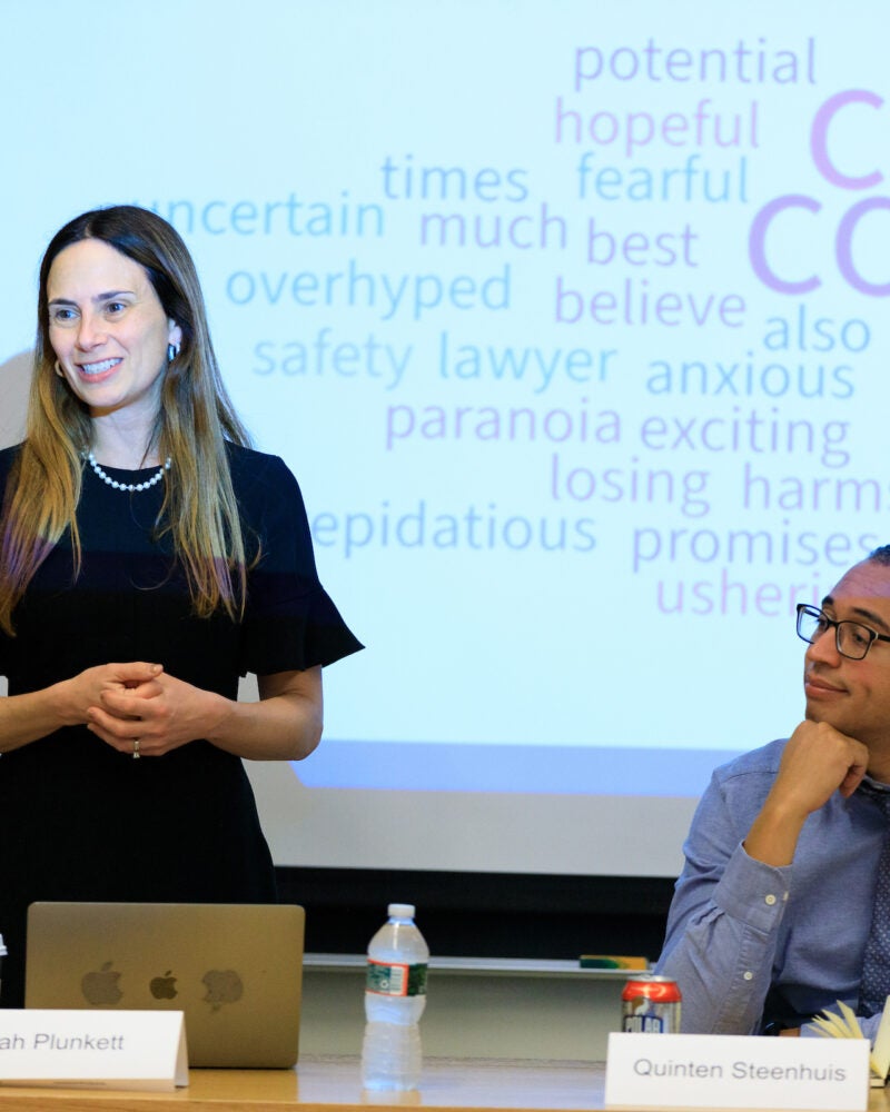 a woman stands in front of a projector screen, a man sits next to her