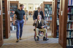 A man in a wheelchair is being pushed through a library as a young man speaks with him.