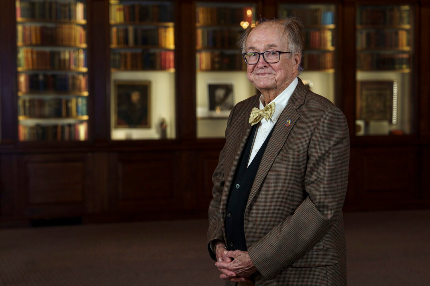 A portrait of a man in a plaid jacket and yellow bow tie in front of a lighted bookcase.