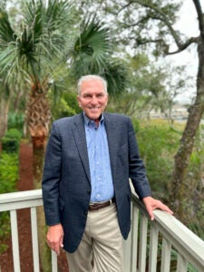 A portrait of a man standing outside on a porch with greenery behind him