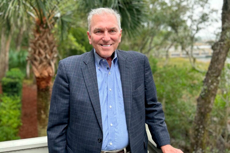 A portrait of a man standing outside on a porch with greenery behind him