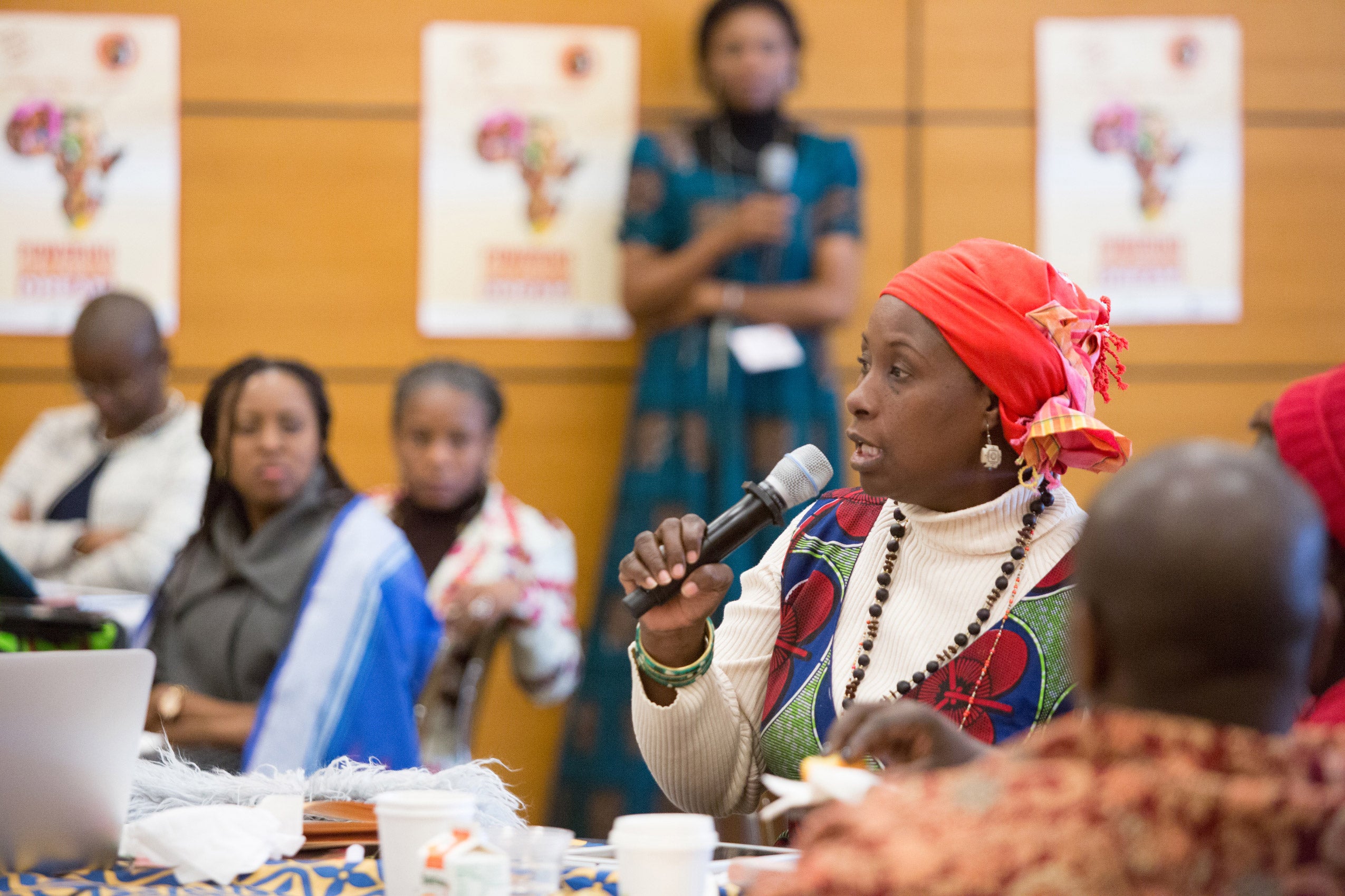 A woman sitting and speaking into a microphone while others look on and listen