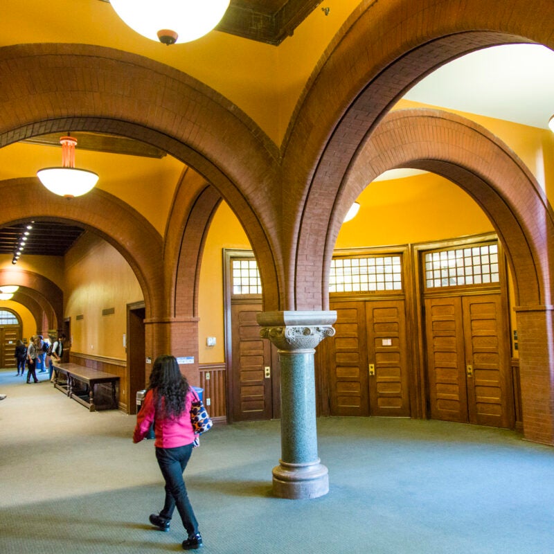 A student walks down the first floor hallway beneath the brick interior archways of a building