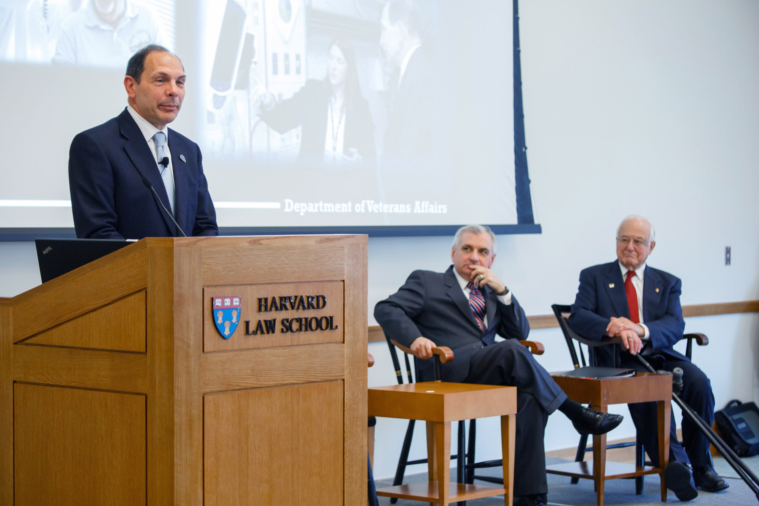 Three men on stage - two seated and one standing and speaking at a podium
