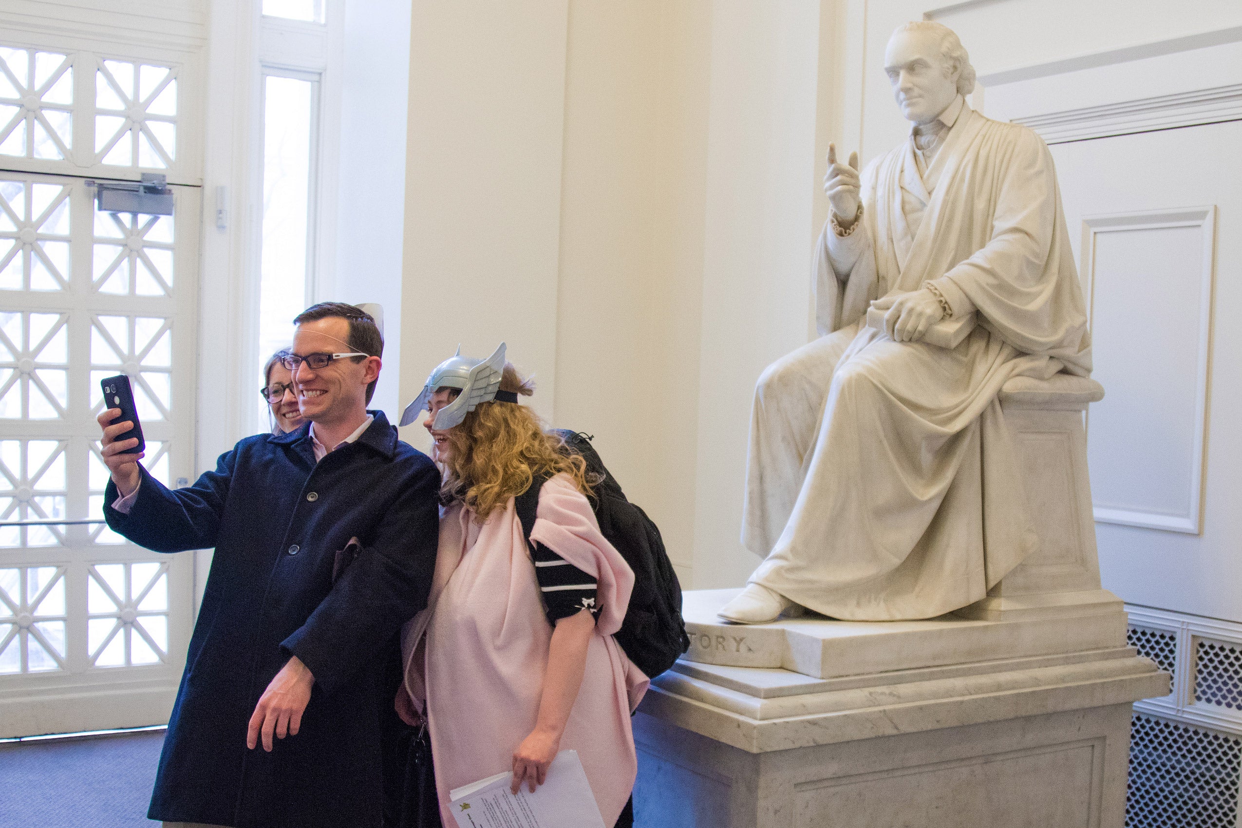 Two women and a man take a selfie with a statue in a lobby of Langdell Hall