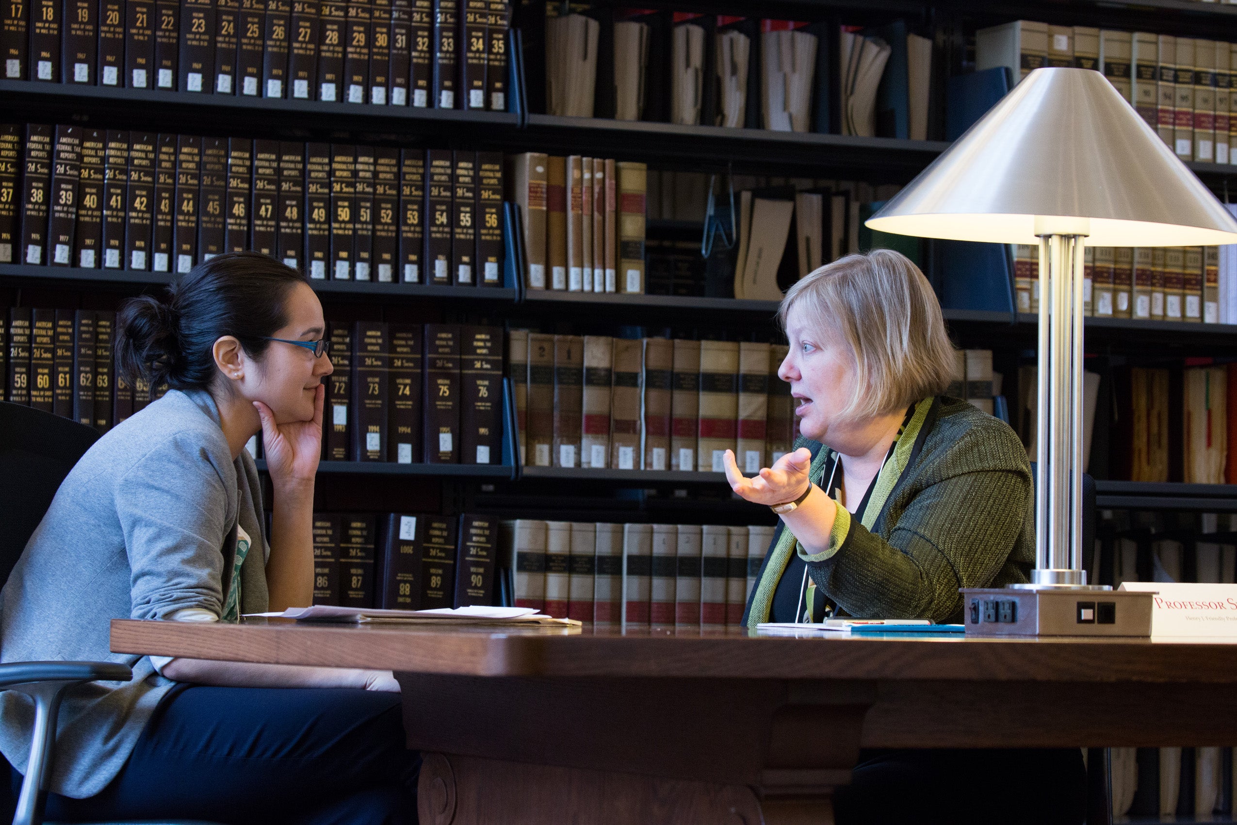 Two women sitting at a table in front of the library stacks having a conversation