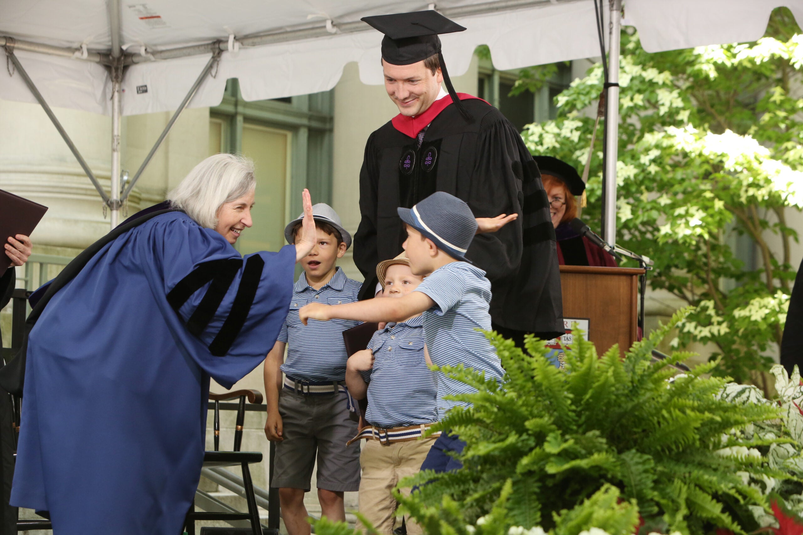 A graduate on stage with three little boys, one of whom is being high fived by a woman