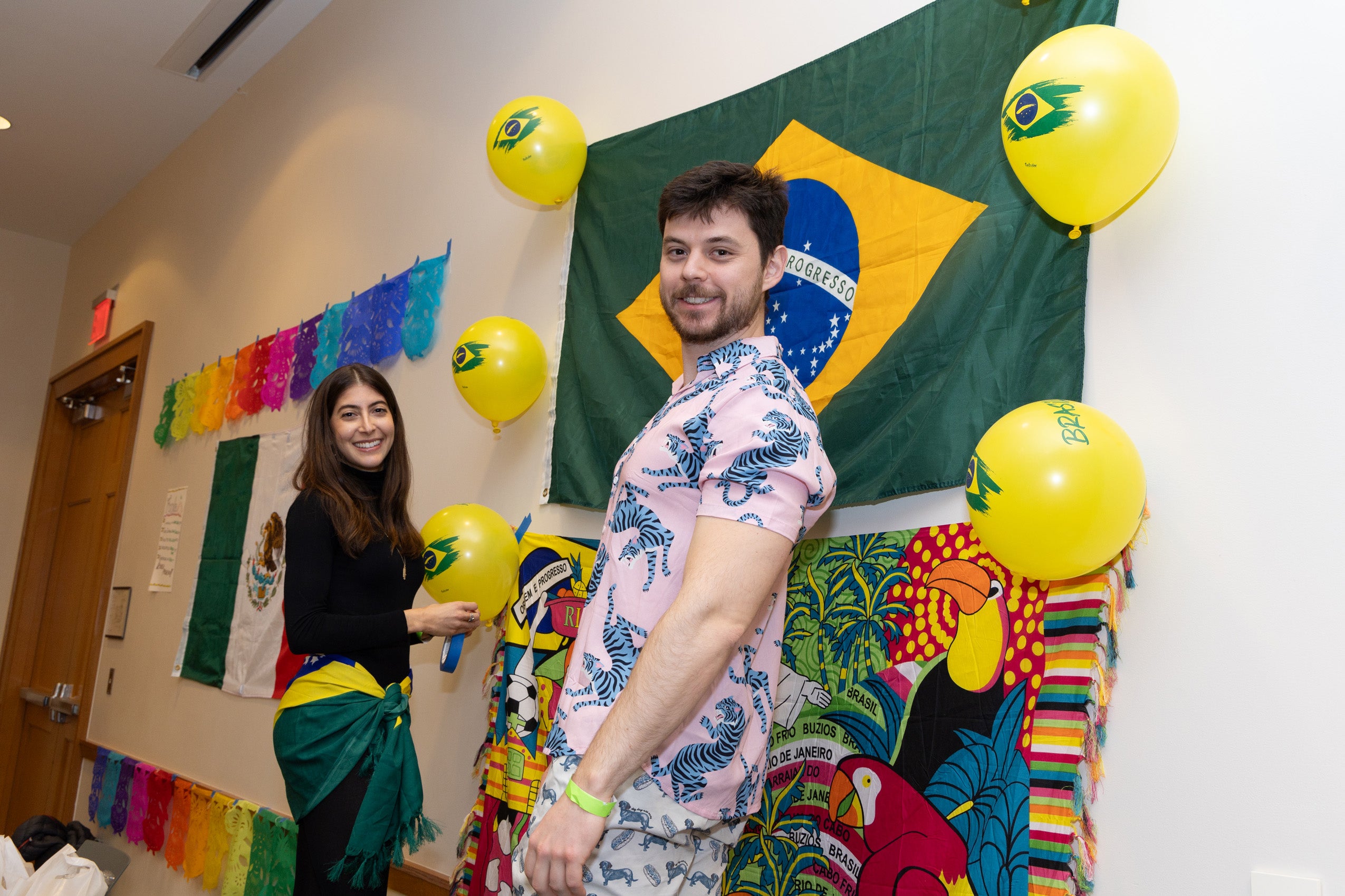 A man wearing a festive shirt and a woman decorate a wall with a green and yellow flag and balloons.