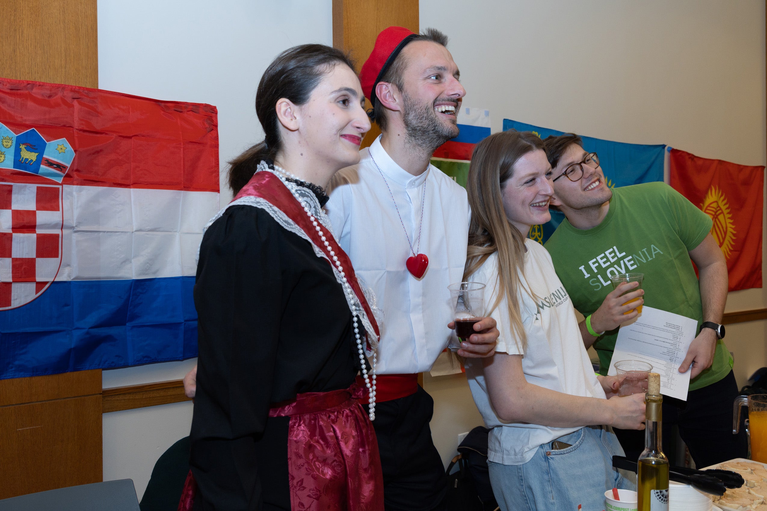 Four students, two wearing clothing from their native country pose in front of an international flag at an event.