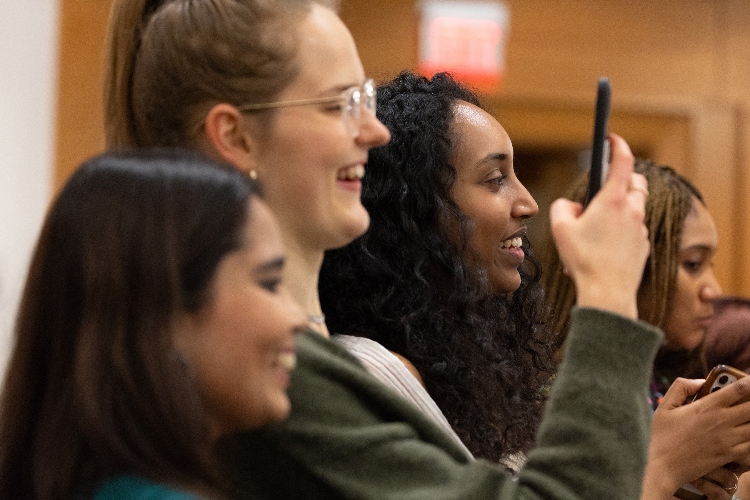 Several women in an audience smile and take pictures with their cell phones.